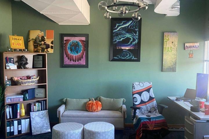 A cozy office with green walls and various wall art including vibrant abstract paintings. A small couch with a pumpkin placed on it is flanked by two round stools. A blanket with a bold design covers a chair near a desk. An open shelving unit displays books and decorative items.