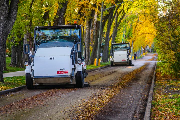 Street sweepers from Elgin are cleaning a tree-lined road covered with fallen leaves. The scene is vibrant, with trees displaying shades of green and yellow along the sidewalk.