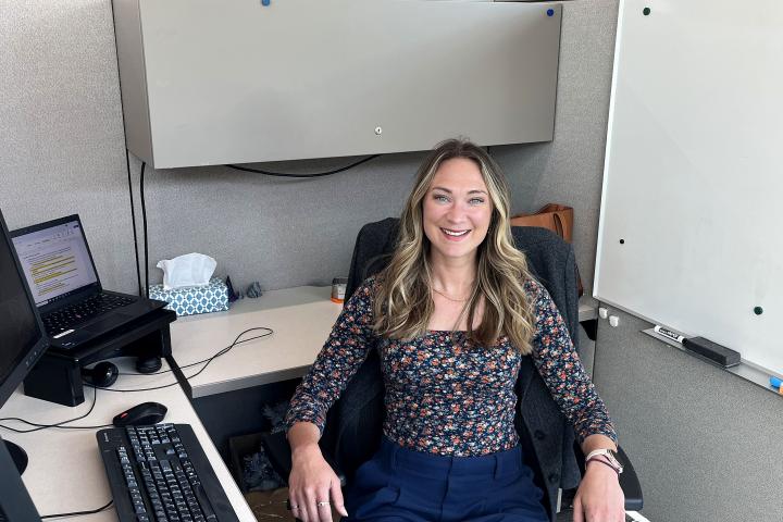 Photo of a woman sitting at a desk in an office environment, posing for a casual yet professional photo.