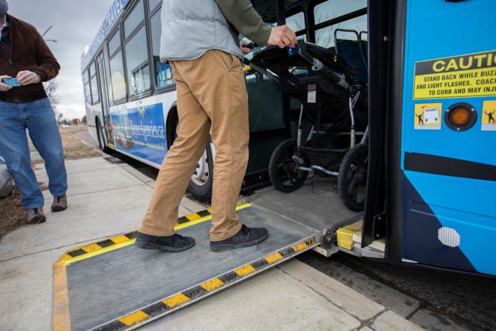 A person is pushing a stroller up a bus ramp from the sidewalk. Another person stands nearby. The bus is painted in blue and white. There is a caution sign near the entrance.