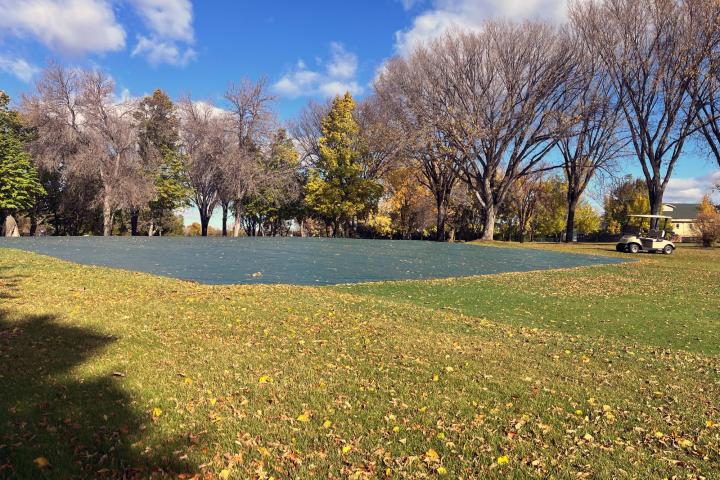 A large outdoor golf course covered with a tarp in a park, surrounded by trees with autumn foliage. A golf cart is parked nearby. The sky is clear with a few scattered clouds.