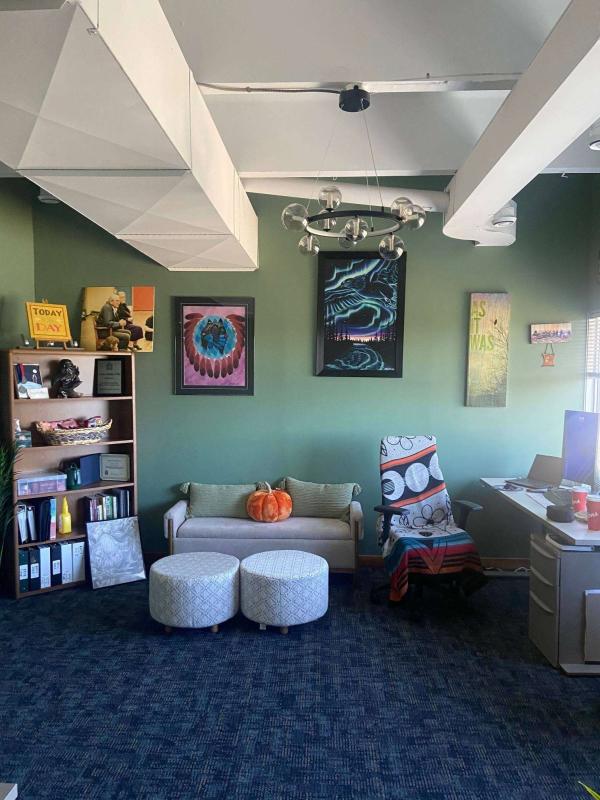 A cozy office with green walls and various wall art including vibrant abstract paintings. A small couch with a pumpkin placed on it is flanked by two round stools. A blanket with a bold design covers a chair near a desk. An open shelving unit displays books and decorative items.