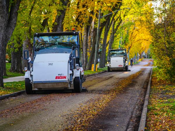 Street sweepers from Elgin are cleaning a tree-lined road covered with fallen leaves. The scene is vibrant, with trees displaying shades of green and yellow along the sidewalk.