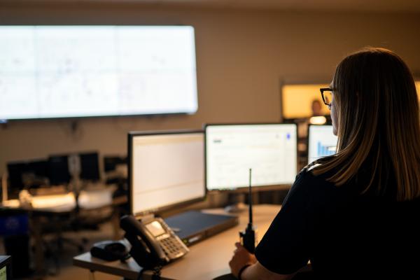 A person is seated at a desk in a control room, monitoring multiple computer screens displaying data and charts. A large wall display with additional information is in the background. The workspace includes a phone and a radio. The room has dim lighting, creating a focused atmosphere.