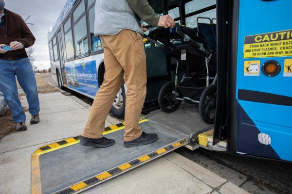 A person is pushing a stroller up a bus ramp from the sidewalk. Another person stands nearby. The bus is painted in blue and white. There is a caution sign near the entrance.