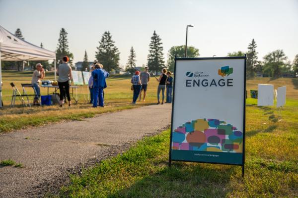 A group of people is gathered in a park near a white tent, with trees in the background. A large sign in the foreground reads "City of Saskatoon Engage" with colorful speech bubbles as a background design.
