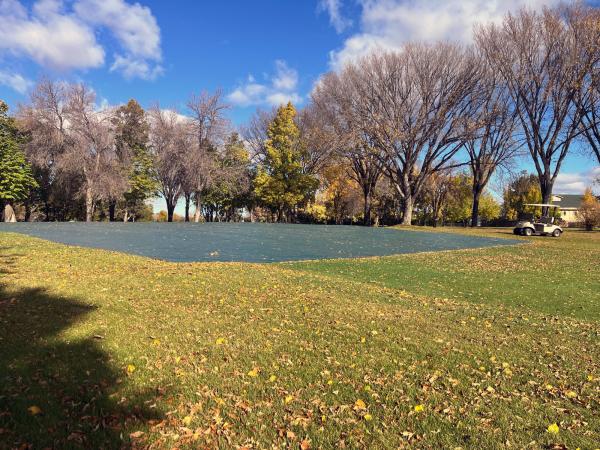 A large outdoor golf course covered with a tarp in a park, surrounded by trees with autumn foliage. A golf cart is parked nearby. The sky is clear with a few scattered clouds.