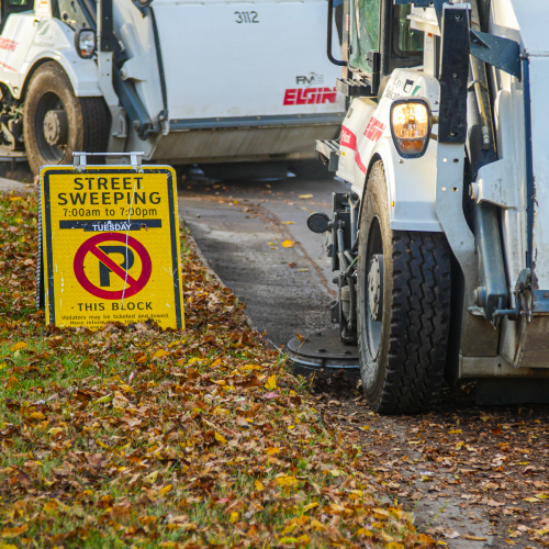A street sweeper vehicle from the brand Elgin is operating near a curb covered with autumn leaves. A yellow sign indicating "Street Sweeping 7:00am to 7:00pm" is placed nearby.