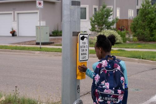 A person wearing a floral backpack pushes a button on a pedestrian crossing signal. The signal is on a roadside with houses and greenery in the background.