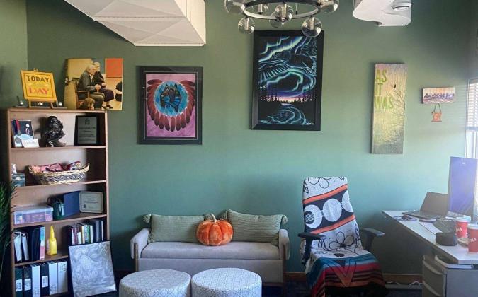 A cozy office with green walls and various wall art including vibrant abstract paintings. A small couch with a pumpkin placed on it is flanked by two round stools. A blanket with a bold design covers a chair near a desk. An open shelving unit displays books and decorative items.