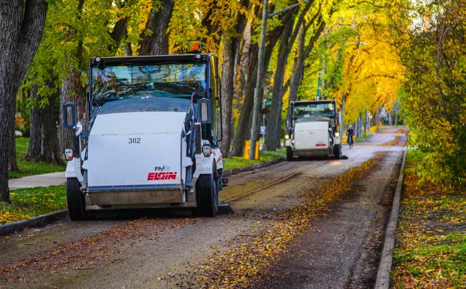Street sweepers from Elgin are cleaning a tree-lined road covered with fallen leaves. The scene is vibrant, with trees displaying shades of green and yellow along the sidewalk.