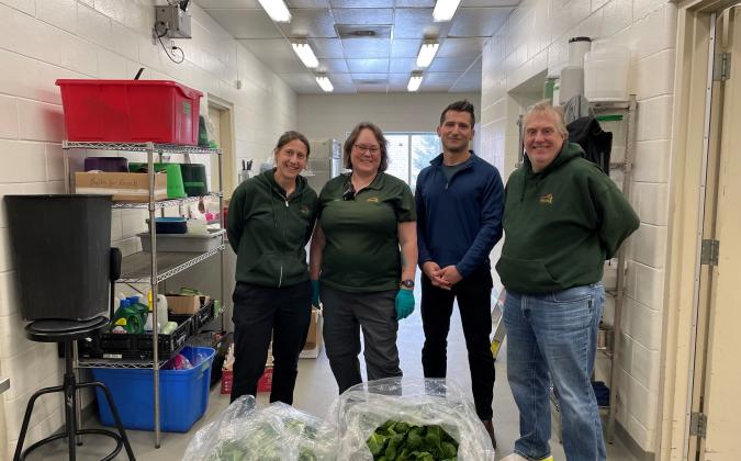 Four people stand in a bright indoor workspace beside large open bags filled with green lettuce.