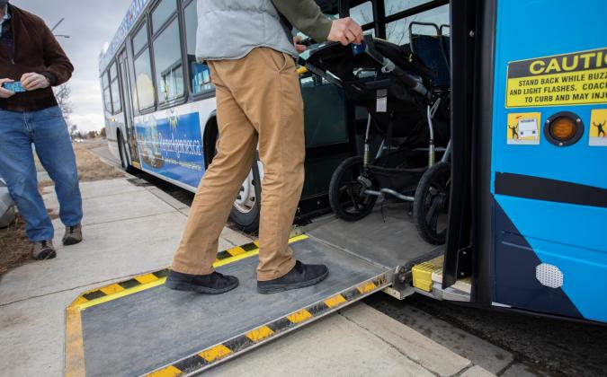A person is pushing a stroller up a bus ramp from the sidewalk. Another person stands nearby. The bus is painted in blue and white. There is a caution sign near the entrance.