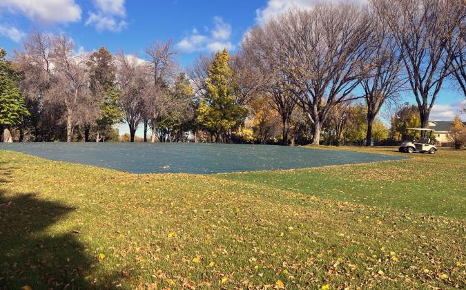 A large outdoor golf course covered with a tarp in a park, surrounded by trees with autumn foliage. A golf cart is parked nearby. The sky is clear with a few scattered clouds.